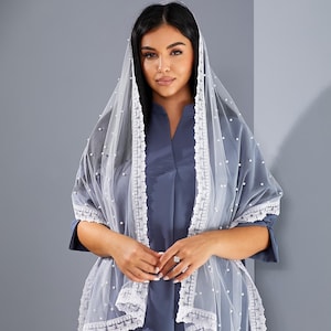 A woman wearing a blue dress and a white veil with lace trim stands against a gray background.