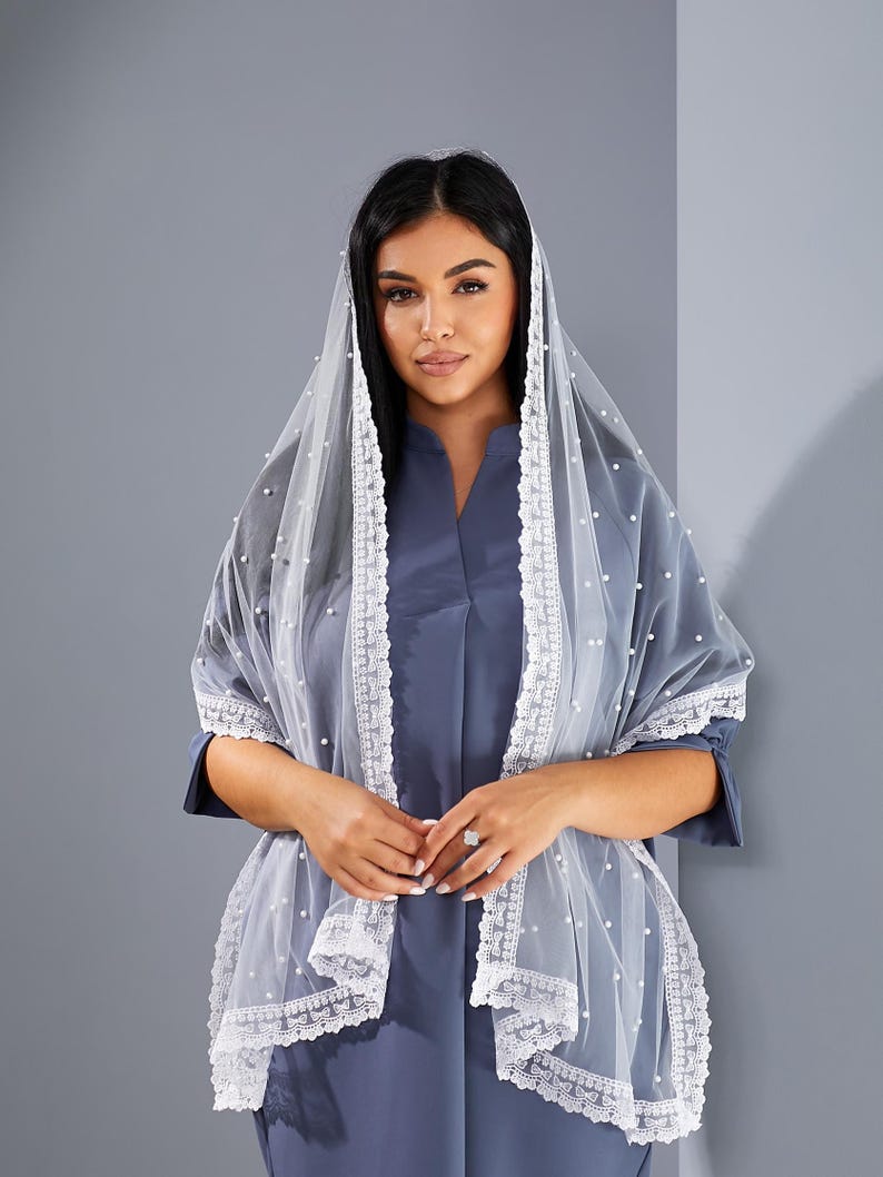 A woman wearing a blue dress and a white veil with lace trim stands against a gray background.