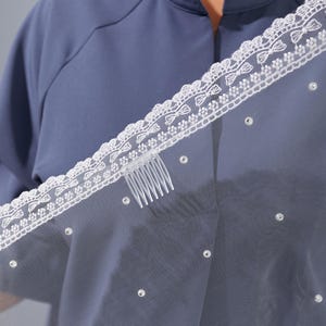 A close-up view of a person wearing a blue shirt with a white lace-trimmed veil draped over their head.