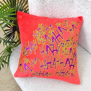 May include: A square, coral-colored decorative pillow with the word "HA HA HA" in a repeating pattern. The text is in purple with a green outline. The pillow is on a white, textured surface.