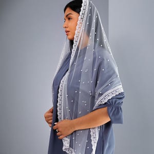 A woman wearing a blue dress with a white lace veil stands against a gray background.