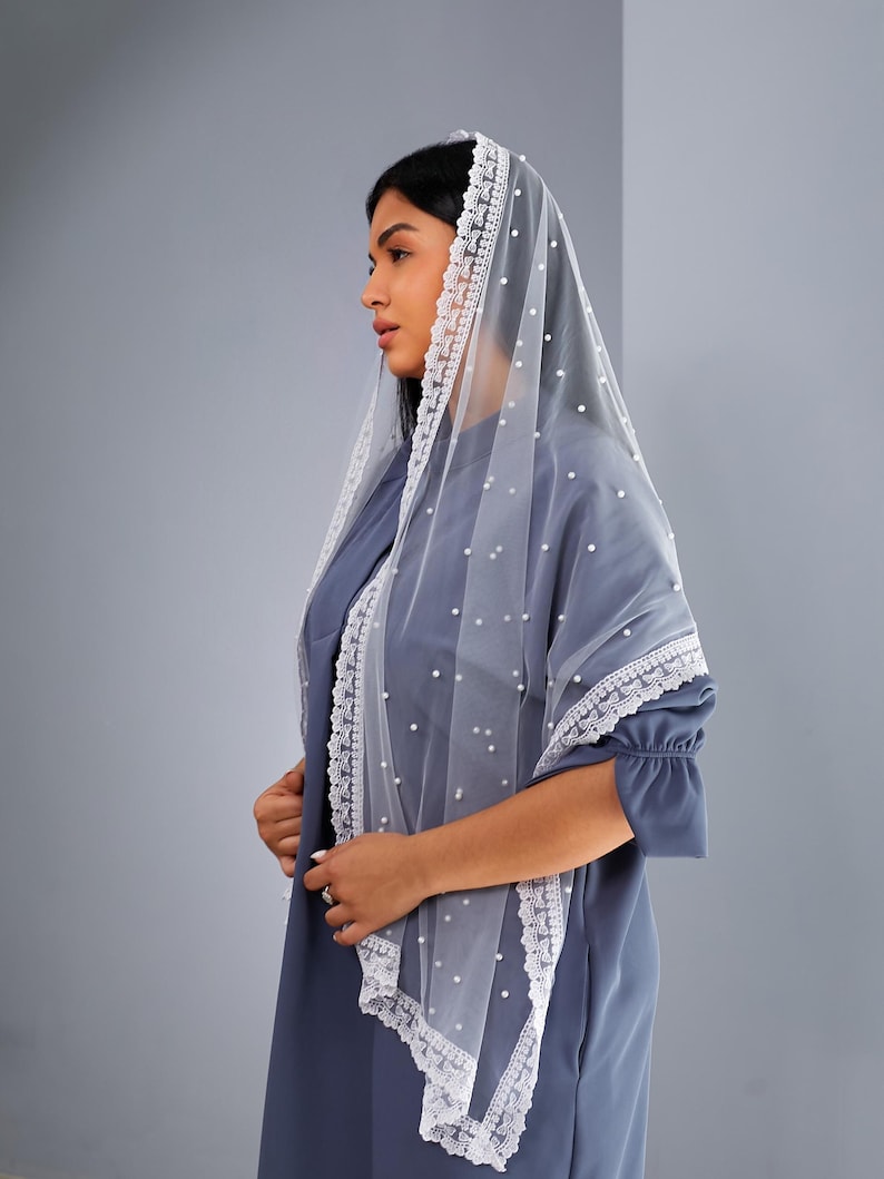 A woman wearing a blue dress with a white lace veil stands against a gray background.