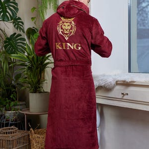 以下が含まれることがあります： ゴールドのライオンのエンブレムと背中に「KING」の文字が刺繍された、バーガンディ色のフード付きローブ。 ローブにはベルトが付いており、柔らかく豪華な素材で作られています。 ローブは膝丈です。