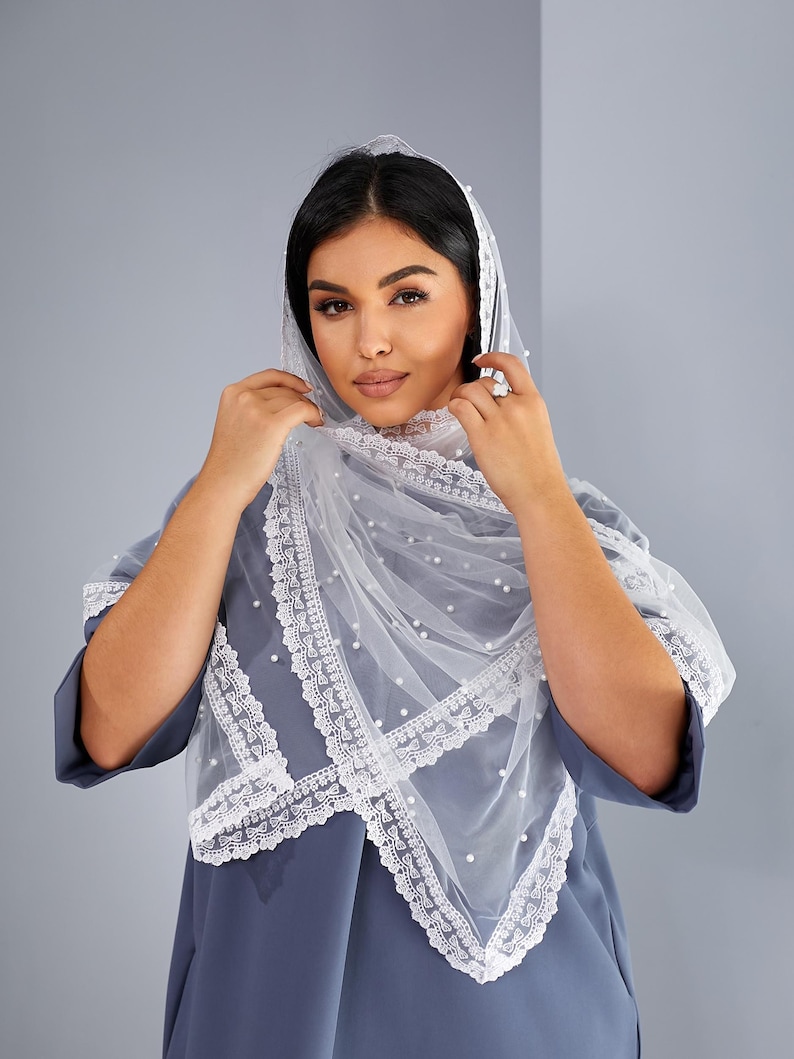 A woman wearing a white veil with lace trim and a gray dress stands against a gray background, adjusting her veil.