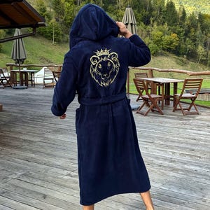 Op de afbeelding: Marineblauwe badjas met capuchon en een gouden leeuwenkamontwerp op de rug. De badjas heeft een riem en zakken. De persoon staat op een houten terras met een schilderachtige achtergrond van bomen en een meer.