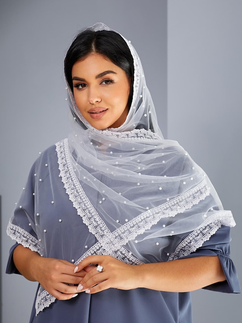 A woman wearing a white veil with lace trim and a gray dress stands against a gray background, smiling at the camera.