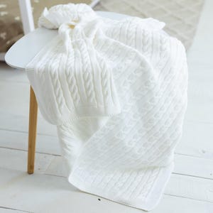 May include: A soft white knitted blanket featuring a classic cable knit pattern with vertical braided textures. The fabric has a clean, uniform appearance with a ribbed border edge, draped over a surface to showcase the intricate woven design.
