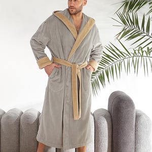 May include: A grey hooded bathrobe with tan trim and a matching belt. The robe has pockets and is made of a soft, plush material. The model is standing on a cream-coloured rug.