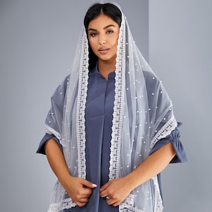 A woman wearing a blue dress and a white veil with lace trim stands against a gray background.