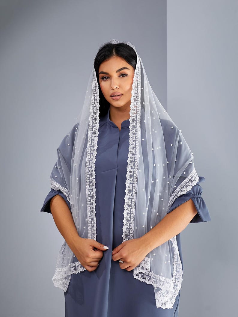 A woman wearing a blue dress and a white veil with lace trim stands against a gray background.