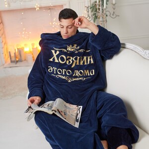 May include: A navy blue hooded blanket with gold text that reads "Хозяин этого дома". A person is wearing the blanket and sitting on a white sofa, holding a magazine. The room is decorated with Christmas lights and candles.