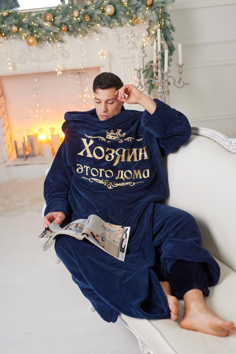 May include: A navy blue hooded blanket with gold text that reads "Хозяин этого дома". A person is wearing the blanket and sitting on a white sofa, holding a magazine. The room is decorated with Christmas lights and candles.