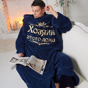 May include: A navy blue hooded robe with gold text that reads "Хозяин этого дома" (Master of this house). The person is sitting on a white couch and holding a magazine. The robe is made of a soft, plush material.