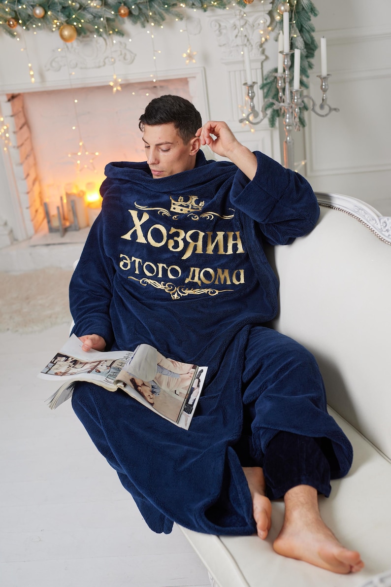May include: A navy blue hooded robe with gold text that reads "Хозяин этого дома" (Master of this house). The person is sitting on a white couch and holding a magazine. The robe is made of a soft, plush material.