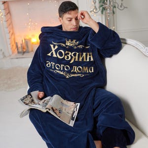 May include: A navy blue hooded blanket with gold text reading "Хозяин этого дома". A person is wearing the blanket and sitting on a white sofa, holding a magazine. The room is decorated with festive lights and candles.