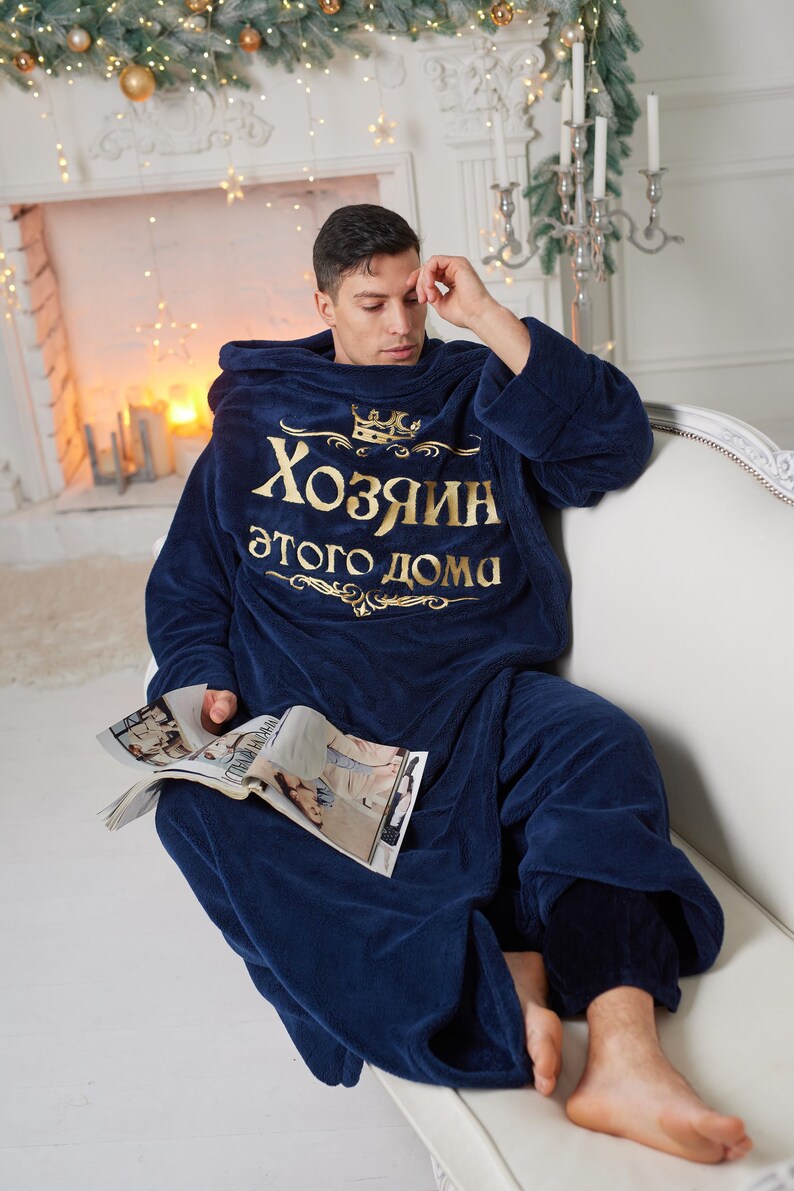 May include: A navy blue hooded blanket with gold text reading "Хозяин этого дома". A person is wearing the blanket and sitting on a white sofa, holding a magazine. The room is decorated with festive lights and candles.