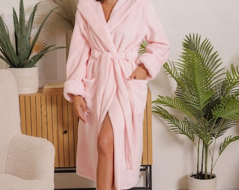 May include: A light pink hooded bathrobe with a matching belt and pockets. The plush robe is knee-length and has long sleeves. The person is standing in a well-lit room with a wooden cabinet, a white chair, and potted plants.