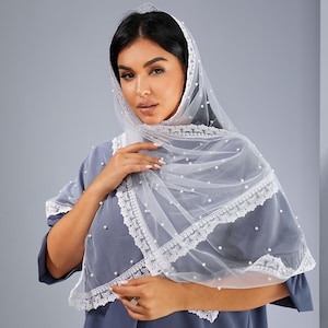 A woman wearing a white veil with lace trim and a gray dress stands against a gray background.
