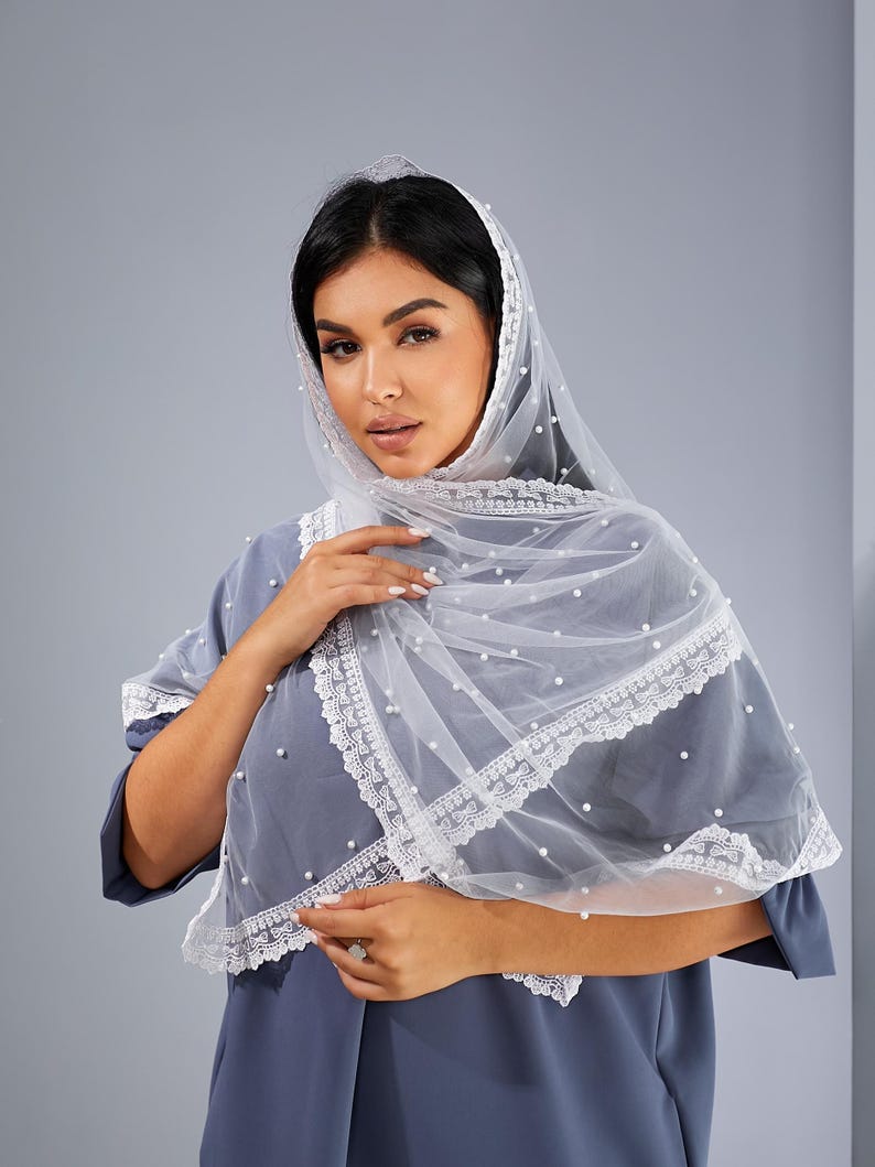 A woman wearing a white veil with lace trim and a gray dress stands against a gray background.