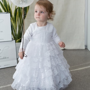 May include: A young child wearing a white, long-sleeved dress with multiple layers of lace. The dress has a round neckline and lace detailing on the sleeves. The child is holding a clear balloon with white specks.