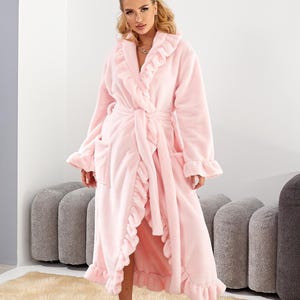 May include: A light pink, plush robe with ruffled trim and a matching belt. The robe has long sleeves and pockets. The model is standing on a beige rug.