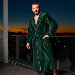 May include: A dark green velvet robe with a hood and a matching belt. The robe has gold trim along the edges and pockets. The person is standing on a balcony with a sunset in the background.