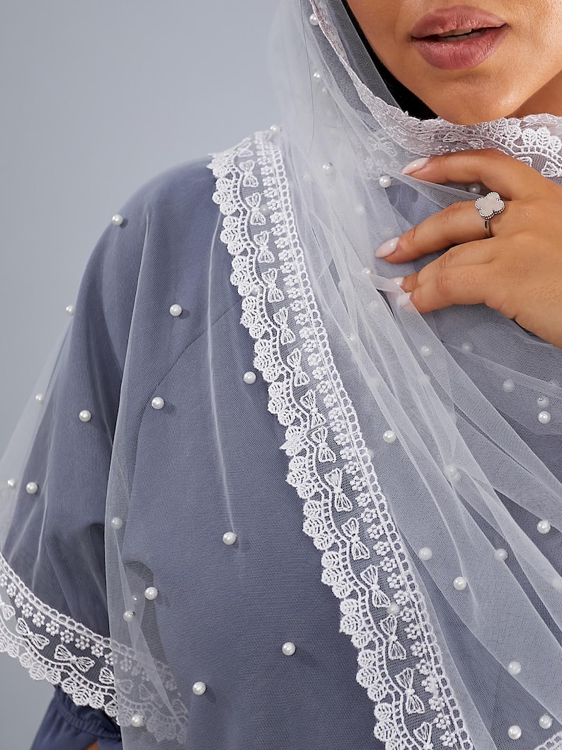 A close-up of a person wearing a gray dress with a white lace-trimmed veil and a ring on their finger.