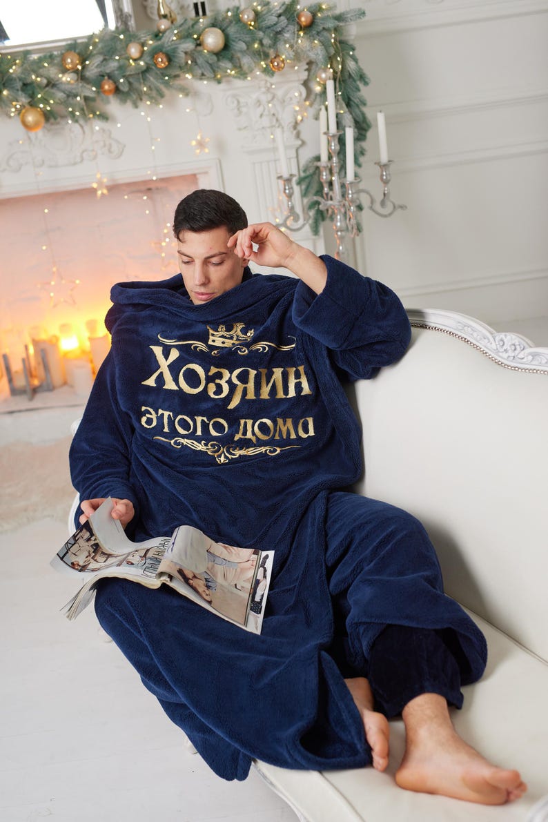 May include: A navy blue hooded blanket with gold text reading "Хозяин этого дома" (Master of this house). A person is wearing the blanket and reading a magazine on a white sofa. The room is decorated with Christmas lights and a fireplace.