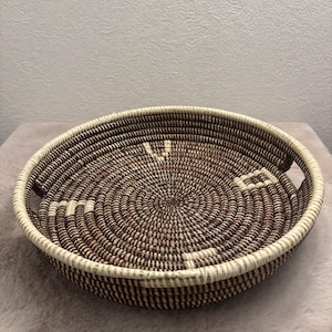 Peut inclure: Un panier rond et peu profond en fibres tressées, présentant un motif en spirale alternant des tons brun foncé et crème. Le panier possède un bord épais renforcé et deux poignées latérales intégrées, avec des motifs géométriques crème.