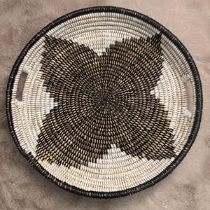 May include: A round, woven basket tray featuring a spiral pattern in black, white, and natural tan fibers. The tray has a raised rim and two integrated side handles, with the woven design radiating from the center toward the outer edge.