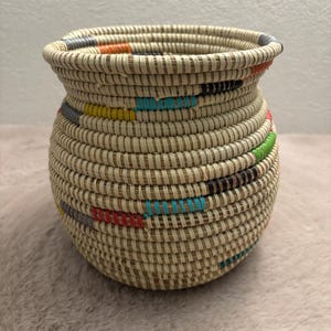 Peut inclure: Un panier rond tissé à la main en fibres naturelles beige clair. Le panier présente un bord large et évasé ainsi qu'un corps bombé décoré de petits accents horizontaux de fils colorés dans des tons orange, jaune, bleu, vert, rouge et noir.