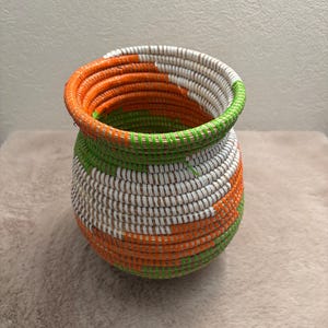 Peut inclure: Un petit panier rond enroulé présentant un motif vibrant de fibres tissées orange, vert vif et blanc. Le panier possède une large ouverture supérieure avec un col légèrement effilé et une base arrondie, fait de matériaux étroitement enroulés.
