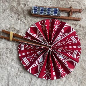 Peut inclure: Éventail rond plié à motifs rouges et blancs avec des poignées en bois et un hochet assorti. Le motif géométrique orne l'éventail. Le hochet présente un motif tissé bleu et blanc.