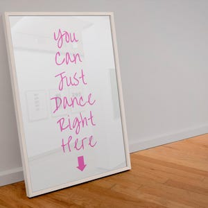 May include: A framed mirror with the text "You Can Just Dance Right Here" in pink script. A pink arrow points down. The mirror is in a white frame and leans against a white wall and wooden floor.