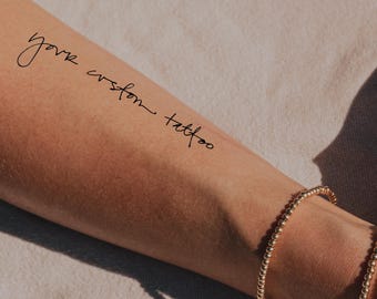 May include: A temporary tattoo on an arm with the words "your custom tattoo" in a handwritten script. A gold beaded bracelet is visible on the wrist. The skin tone is light brown.