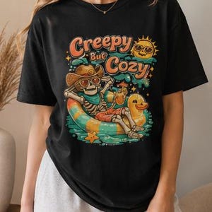 Op de afbeelding: Een zwart t-shirt met korte mouwen en een grafische print van een skelet met een cowboyhoed en zonnebril, ontspannend in een gele eend-zwemband. De tekst "Creepy But Cozy" staat boven het skelet met een zon-icoon.