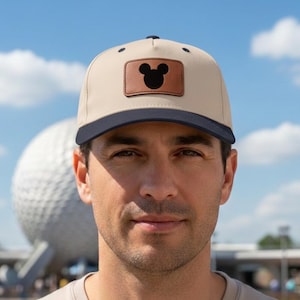 May include: Tan baseball cap with a navy blue brim and a brown leather patch featuring a black Mickey Mouse silhouette. The cap is worn by a person in a light-colored t-shirt, with a blurred background of a large white sphere and blue sky.