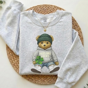 May include: A light gray sweatshirt featuring a teddy bear wearing a green beanie, gray hoodie with "FEELING LUCKY" text, blue jeans, and white sneakers. The bear holds a four-leaf clover. A gold heart pendant necklace is visible.