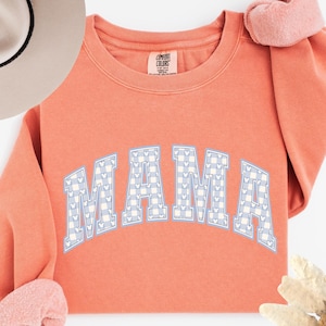 May include: Coral-colored sweatshirt with the word "MAMA" in large, light blue and white gingham letters. The sweatshirt has a crew neck and long sleeves with pink cuffs. A light-colored hat is visible in the upper left corner.