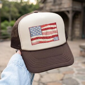 May include: A brown and tan trucker hat with a stitched American flag design. The flag features blue stars, red stripes, and a white background. The hat has a brown brim and mesh back.