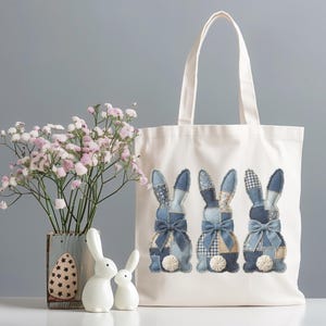 May include: A white tote bag features three denim patchwork bunny designs with blue bows. The bag is next to a vase of pink flowers, two white ceramic bunnies, and a decorative egg. The scene is set on a white surface.