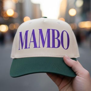 May include: A beige baseball cap with a green brim and the word "MAMBO" in purple lettering. The hat is held up in front of a blurred city background.