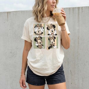 May include: Cream-colored t-shirt featuring a four-panel design of Minnie Mouse in various poses. Each panel showcases Minnie with a leopard print bow and outfit. The background includes vertical stripes in green and tan.