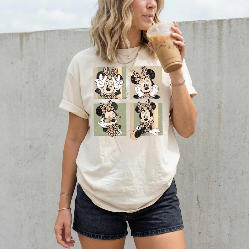 May include: Cream-colored t-shirt featuring a four-panel design of Minnie Mouse in various poses. Each panel showcases Minnie with a leopard print bow and outfit. The background includes vertical stripes in green and tan.
