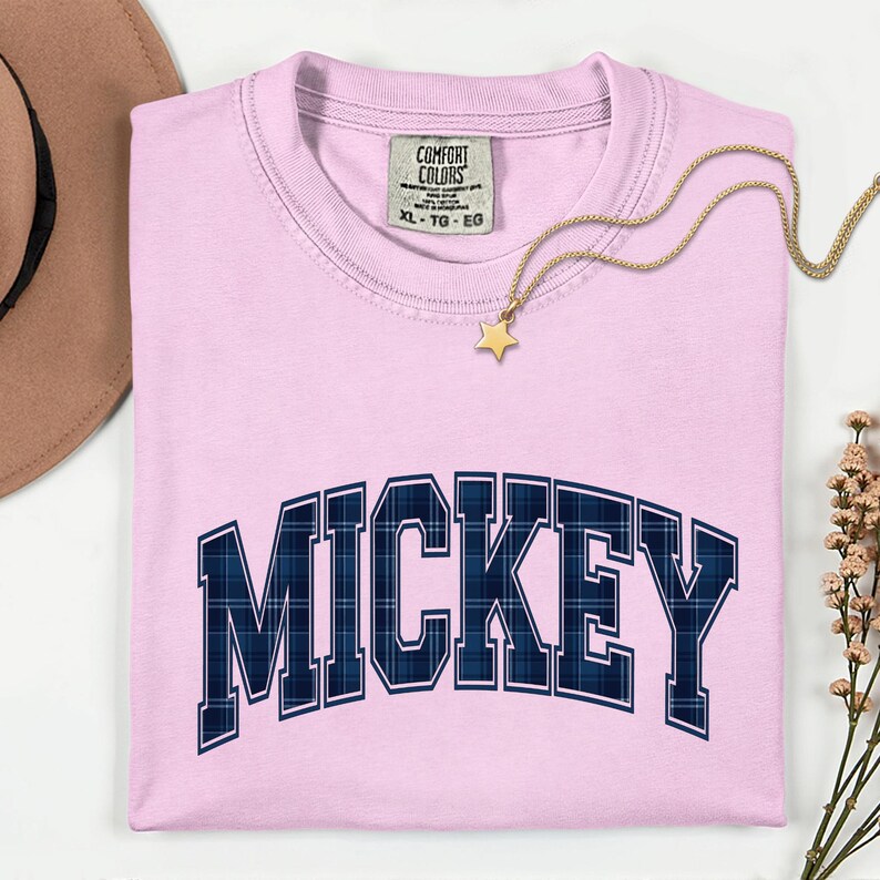 May include: A pale pink t-shirt featuring the word "MICKEY" in navy blue plaid lettering. The shirt has a crew neck and a "COMFORT COLORS" tag. A gold necklace with a star pendant is draped across the shirt.