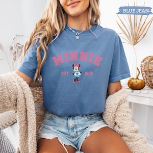 May include: Blue jean-colored t-shirt featuring the name "MINNIE" in pink block letters above a graphic of Minnie Mouse holding a heart. The text "EST 1928" is below the graphic. The shirt has short sleeves and a crew neck.