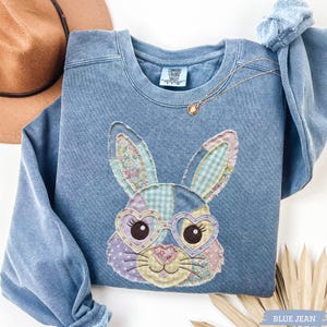 May include: Blue jean-colored sweatshirt with a patchwork bunny face design. The bunny has heart-shaped glasses and is made of various patterned fabrics. A gold necklace and a brown hat are also visible.