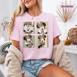 May include: A light pink t-shirt featuring a four-panel graphic of Minnie Mouse in various poses, each with a leopard print bow and outfit. The word "BLOSSOM" is printed in a pink banner at the top. The shirt is worn by a person.
