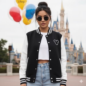 May include: Black and white varsity jacket with a Mickey Mouse outline on the chest. The jacket has white sleeves, a black body, and white stripes on the collar, cuffs, and hem. The background features colorful balloons and a castle.