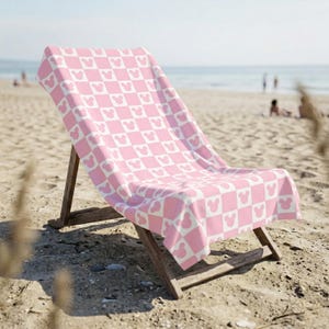 Puede incluir: Una toalla de playa con un patr&oacute;n de tablero de ajedrez repetitivo en rosa claro y blanco, con un peque&ntilde;o icono de silueta de cabeza de rat&oacute;n centrado dentro de cada cuadrado individual de la cuadr&iacute;cula, sobre una silla de playa de madera.